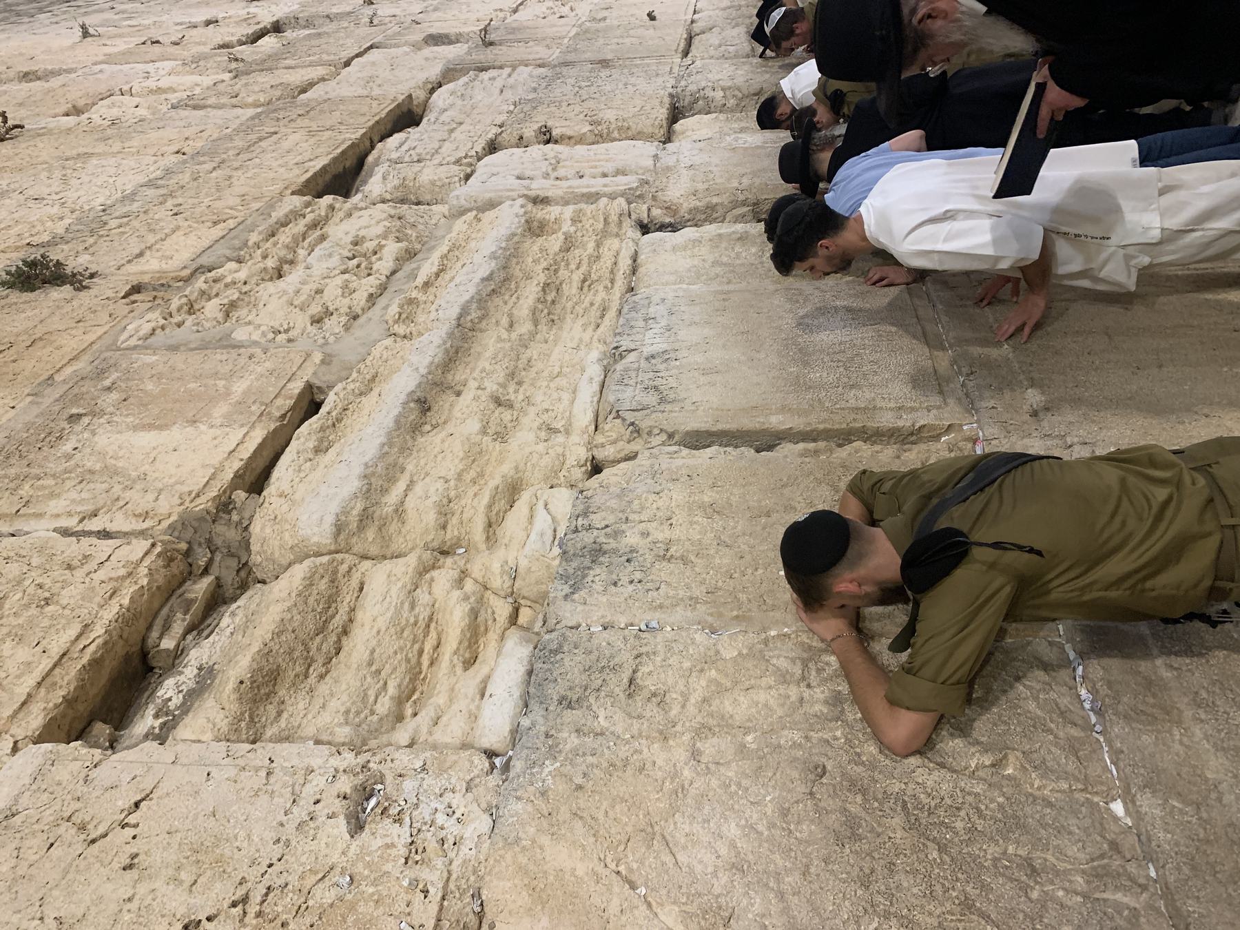 Faces and open palms pressed against the great Herodian stones of the Western Wall at night, a soldier with his forehead on the rock, a man in a white kittel with both hands flat, and folded paper notes in every seam.