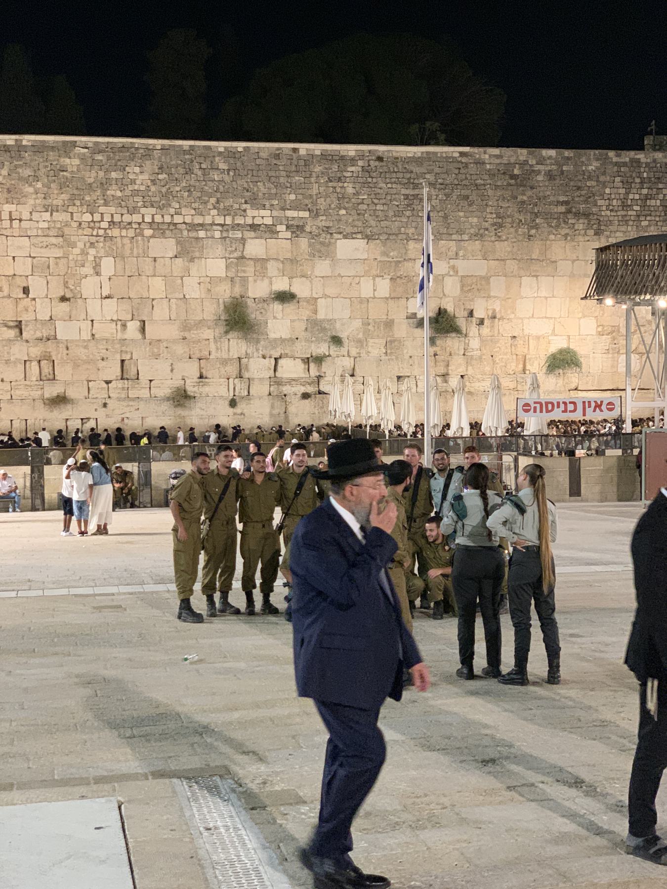 The Western Wall plaza after midnight, floodlit stone rising into a black sky, an Israeli flag on its pole, soldiers and worshippers and an elderly rabbi crossing the open square.