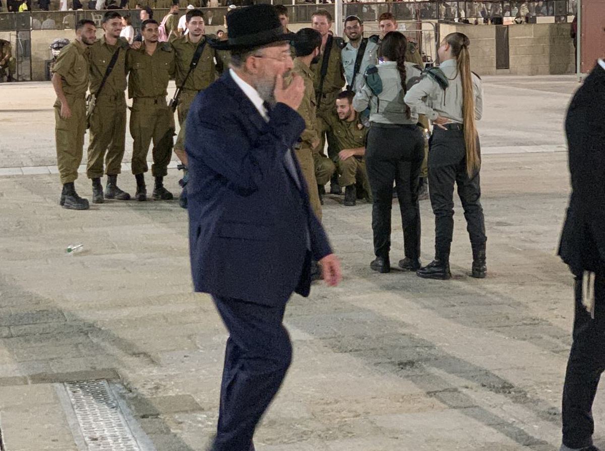An elderly rabbi in a navy suit walking across the plaza past a line of soldiers.