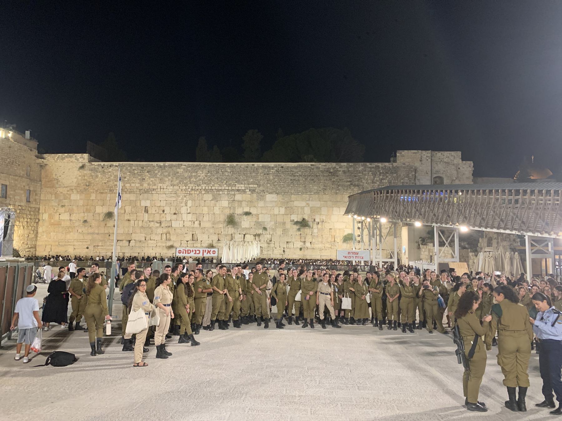 Around two hundred young Israeli soldiers, women and men in olive uniform, standing together in the Western Wall plaza at night facing the lit stones.