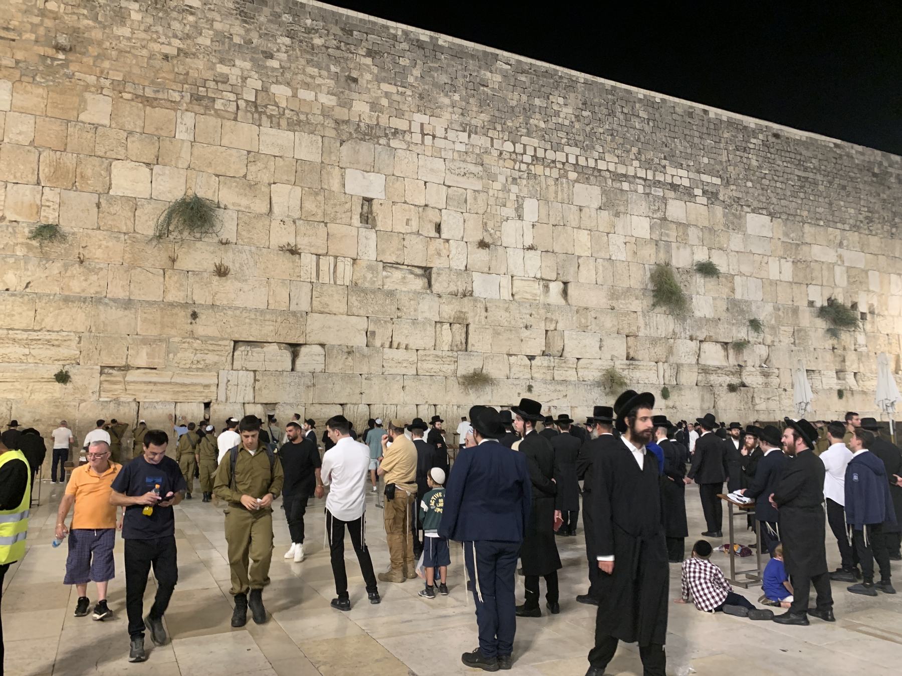 Close along the Western Wall at night, courses of Herodian stone running away in perspective, with a mixed crowd of soldiers, hasidim, families and barefoot children at its base.
